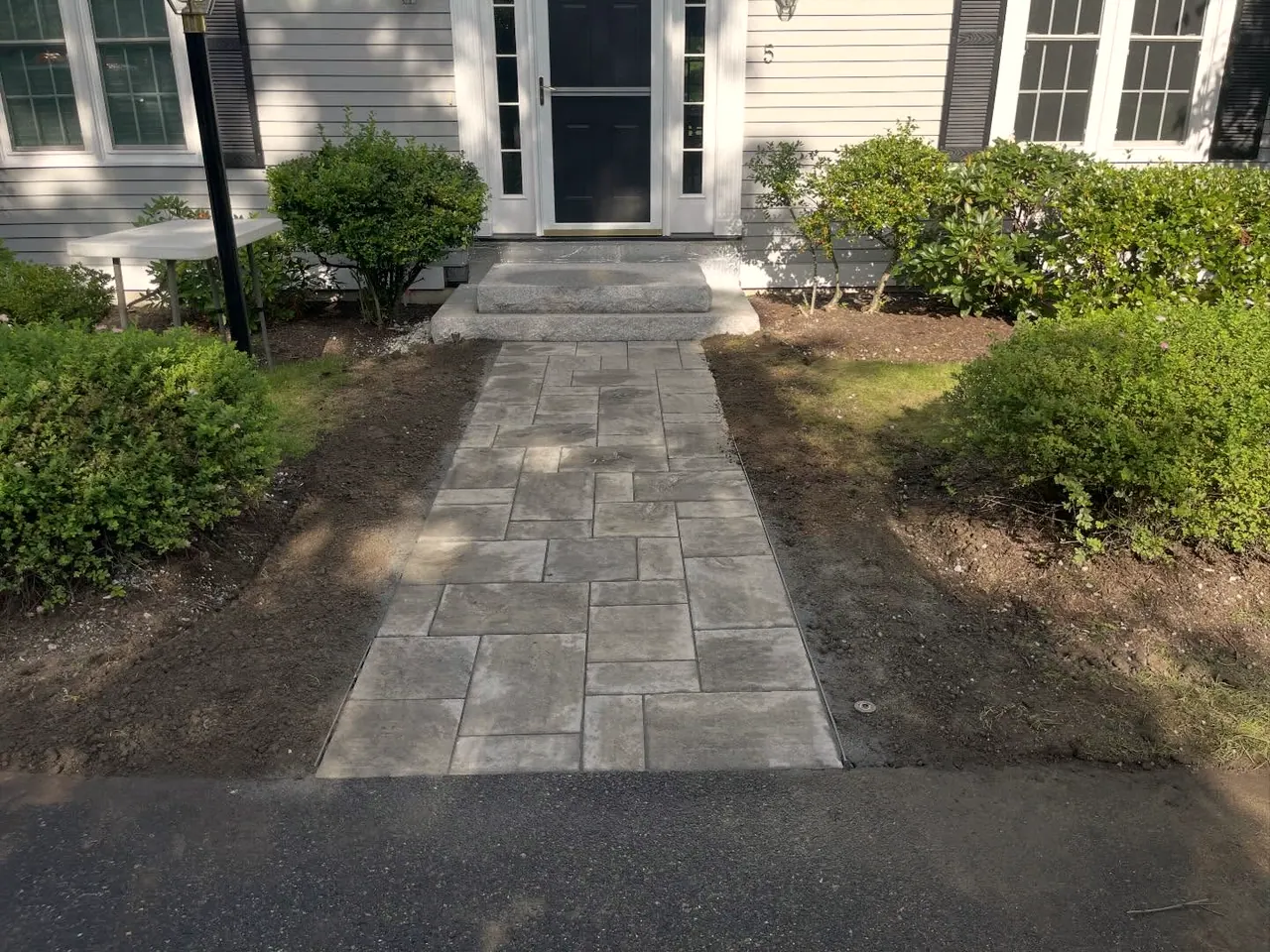 New gray paver walkway leading from the driveway to a front door with a granite step, trimmed shrubs on both sides